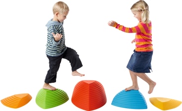 Zwei kleine Kinder balancieren auf großen, bunten, kuppelförmigen Trittsteinen, die in einer Reihe angeordnet sind. Der Hintergrund ist einfarbig weiß. Der Junge trägt einen gestreiften Kapuzenpulli und kurze Hosen, das Mädchen ein gestreiftes Oberteil und einen Jeansrock.