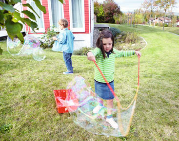 Zwei Kinder spielen mit großen Seifenblasen auf einer Wiese vor einem Haus. Das Mädchen im Vordergrund hält zwei Stöcke mit einer Schnur und erzeugt so eine große Seifenblase, während der Junge im Hintergrund ebenfalls Seifenblasen macht.