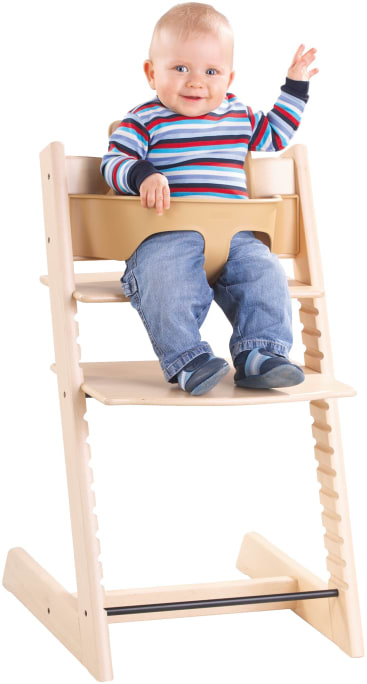 Ein lächelndes Baby in gestreiftem Hemd und Jeans sitzt in einem Holzhochstuhl mit Sicherheitsbügel und hebt eine Hand.