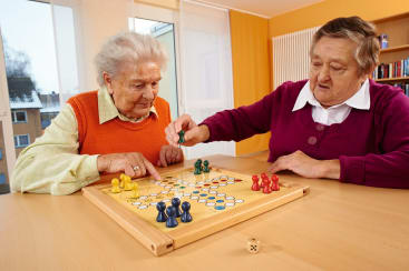 Zwei ältere Frauen sitzen an einem Tisch und spielen ein Brettspiel mit farbigen Steinen und einem Würfel. Sie konzentrieren sich auf ihre Züge in einem hellen, sonnenbeschienenen Raum mit großen Fenstern und Regalen im Hintergrund.
