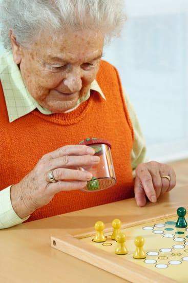 Eine ältere Person in einer orangefarbenen Pulloverweste schüttelt einen Behälter mit Würfeln, während sie auf einem Holztisch ein Brettspiel mit gelben und grünen Spielsteinen spielt.