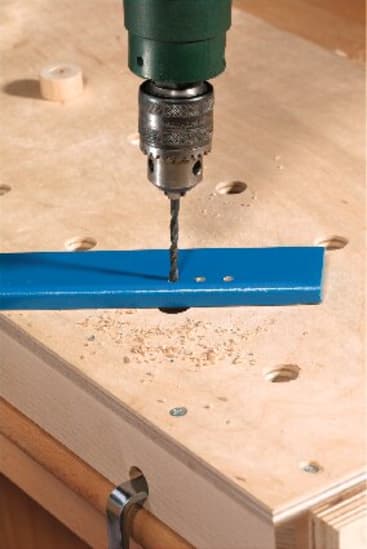 Eine Bohrmaschine bohrt ein Loch in einen blauen Metallstreifen, der auf einer hölzernen Werkbank liegt, wobei Holzspäne um das zu bohrende Loch verstreut sind.