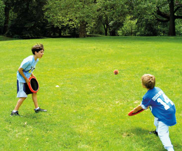 Zwei Kinder spielen mit roten Plastikscheiben und einem kleinen Ball auf einer Wiese Fangen. Beide sind leger gekleidet; ein Kind trägt ein blaues Hemd mit der Nummer 14 auf dem Rücken. Im Hintergrund sind Bäume und Grünflächen zu sehen.