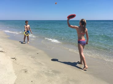 Zwei Kinder in Badeanzügen spielen mit runden Paddeln und einem kleinen Ball an einem Sandstrand in der Nähe der Küste unter einem klaren blauen Himmel Fangen.