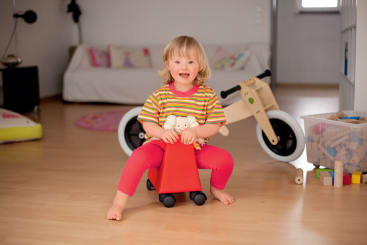 Ein kleines Kind mit hellem Haar sitzt auf einem kleinen roten Spielzeug und hält ein Stofftier. Dahinter stehen ein weißes Laufrad, ein durchsichtiger Spielzeugeimer und ein Sofa in einem aufgeräumten, hellen Wohnzimmer mit Holzboden.