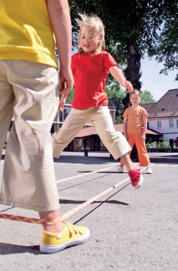 Drei Kinder spielen im Freien ein Springspiel mit einem langen Gummiband. Ein Kind springt gerade über das Band, ein anderes steht innerhalb des Gummibandes und ein drittes Kind schaut im Hintergrund zu.