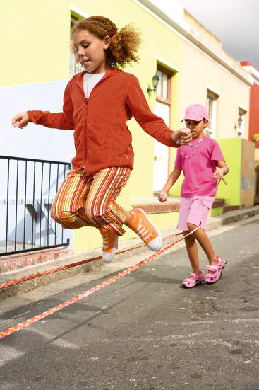 Zwei Kinder spielen draußen auf einer Straße Seilspringen. Ein Kind, das eine orangefarbene Jacke und eine gestreifte Hose trägt, springt, während das andere, das in Rosa gekleidet ist, das Seil dreht. Im Hintergrund sind bunte Gebäude zu sehen.