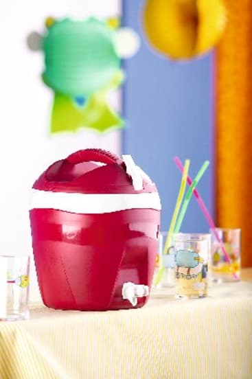 Ein roter Wasserkühler mit weißem Deckel und Ausguss steht auf einer gelb gestreiften Tischdecke. Mehrere klare Gläser mit bunten Motiven und Strohhalmen stehen in der Nähe. Im Hintergrund sind unscharfe, bunte Dekorationen zu sehen.