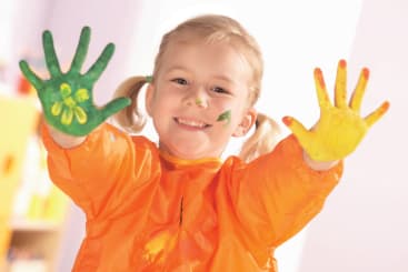 Ein kleines Kind in einem orangefarbenen Kittel lächelt und hält beide Hände hoch, die mit grüner und gelber Farbe bedeckt sind. Es hat einen kleinen Farbklecks im Gesicht. Der Hintergrund ist hell und unscharf.