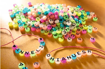Bunte Buchstabenperlen sind auf einer Fläche verstreut, wobei zwei Perlenketten im Vordergrund die Buchstaben "ANNELIE" und "DHIANUESR" darstellen. Einige lose Buchstabenperlen liegen in der Nähe und werden von warmem Licht angestrahlt.