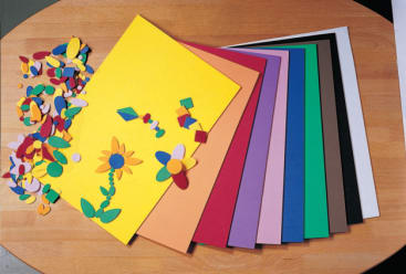 Neun Bögen bunten Bastelschaums sind auf einem Holztisch ausgebreitet, wobei verschiedene Schaumstoffformen, darunter Blumen und geometrische Teile, links von den Bögen angeordnet und verstreut sind.