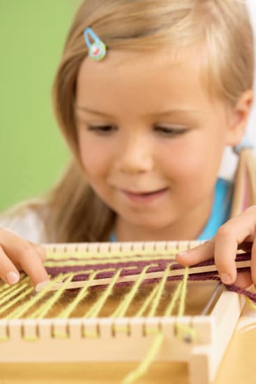 Ein kleines Kind mit blondem Haar webt buntes Garn auf einem kleinen hölzernen Webstuhl, wobei es sich auf das Auffädeln von lila und gelben Fäden konzentriert.