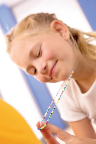 Ein junges Mädchen lächelt und schaut auf eine bunte Perlenkette, die sie mit beiden Händen hält. Sie trägt ein weißes Hemd, und ihr blondes Haar ist zurückgesteckt. Der Hintergrund ist unscharf.