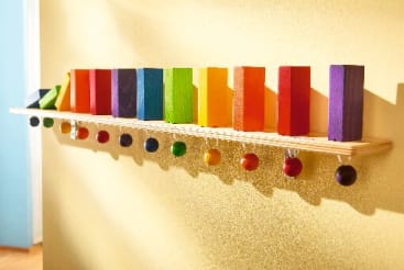 Ein Holzregal, das an einer gelben Wand befestigt ist, enthält aufrecht stehende rechteckige Blöcke in einem Regenbogenfarbverlauf, unter dem Regal hängen farblich passende Kugeln.