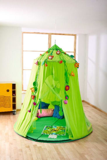 Ein grünes Spielzelt mit Blumen- und Schmetterlingsdekoration steht in einem hellen Raum mit Holzboden. Im Inneren des Zeltes befinden sich mehrere bunte Kissen und ein aufgeschlagenes Bilderbuch. In der Nähe steht ein gelber Schrank.