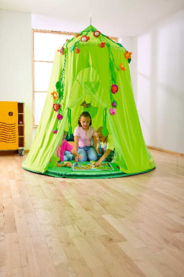Drei Kinder sitzen auf dem Boden in einem grünen Spielzelt und spielen ein Brettspiel. Das Zelt ist mit Kunstblumen geschmückt und hängt von der Decke in einem hellen, spärlich eingerichteten Raum mit Holzboden.