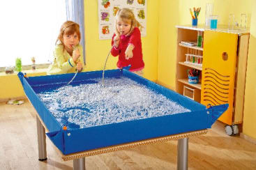 Zwei Kinder stehen an einem blauen Wassertisch, der mit sprudelndem Wasser gefüllt ist. Ein Kind spricht in ein flexibles Rohr, während das andere Kind auf das Wasser zeigt. Der Raum hat gelbe Wände und einen Holzfußboden.