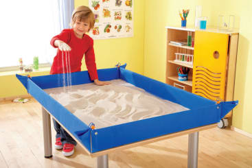 Ein kleines Kind in einem roten Hemd steht an einem mit Sand gefüllten Tisch und lässt den Sand aus einer Hand fallen. Der Tisch hat einen blauen Rand, und der Raum hat gelbe Wände und Unterrichtsmaterialien im Hintergrund.
