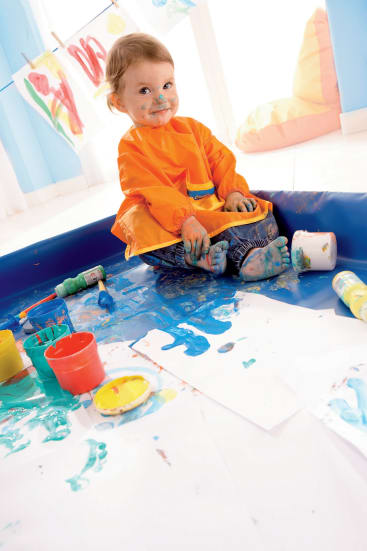 Ein kleines Kind in einem orangefarbenen Kittel sitzt auf einer blauen Matte und malt sich das Gesicht, die Hände und die Füße an. Kunstzubehör und bunte Farbe liegen verstreut herum, und im Hintergrund hängen fertige Gemälde.