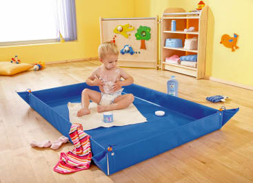 Ein Kleinkind in einer Windel sitzt auf einer weißen Matte in einem blauen Tablett und spielt mit Creme aus einem Glas. Das Zimmer hat gelbe Wände, einen Holzboden und Regale mit Handtüchern und Toilettenartikeln im Hintergrund.