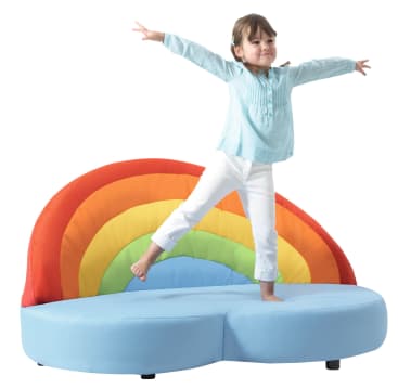 Ein kleines Kind steht mit ausgestreckten Armen auf einem hellblauen wolkenförmigen Sofa mit regenbogenfarbener Rückenlehne vor einem schlichten weißen Hintergrund.