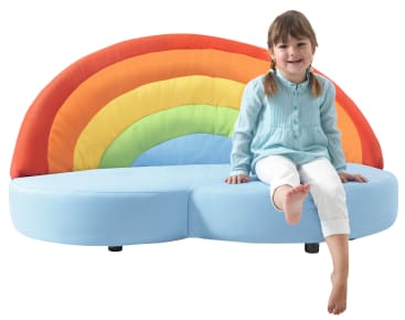 Ein kleines Kind sitzt lächelnd auf einer blauen Couch mit einer regenbogenförmigen, bunten Rückenlehne. Das Kind ist barfuß und trägt ein hellblaues Hemd und eine weiße Hose. Der Hintergrund ist einfarbig und weiß.