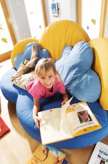 Ein junges Mädchen liegt auf einem blau-gelben wolkenförmigen Sofa, lächelt in die Kamera und hält ein großes Buch aufgeschlagen. Weitere Bücher liegen in der Nähe auf dem Boden. Die Umgebung scheint ein heller Raum zu sein.