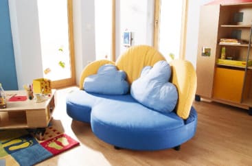 Ein helles Zimmer mit einem blauen und gelben Sofa in Wolkenform, einem niedrigen Tisch mit einem Teeservice, bunten Teppichen, einem Holzboden und Regalen mit Spielzeug und Büchern. Große Fenster lassen viel natürliches Licht herein.