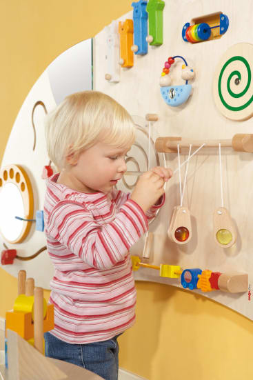 Ein kleines Kind mit blondem Haar und einem rot-weiß gestreiften Hemd interagiert mit einer sensorischen Aktivitätswand, die verschiedene bunte Elemente und Spielzeuge enthält.