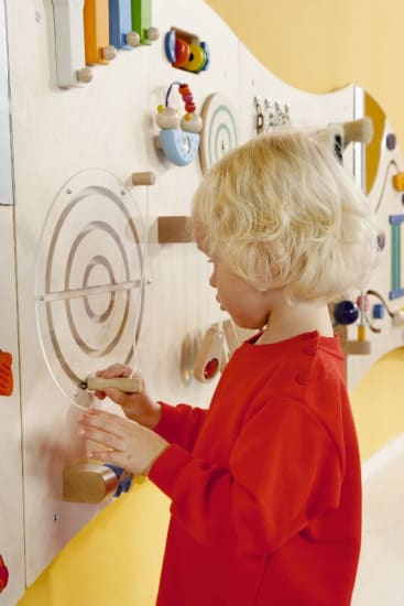 Ein kleines Kind mit blondem Haar und rotem Hemd steht vor einer Wand mit sensorischen Aktivitäten und benutzt seine Hand, um mit einem der kreisförmigen Laborelemente zu interagieren. Die Wand ist mit verschiedenen taktilen Spielzeugen dekoriert.