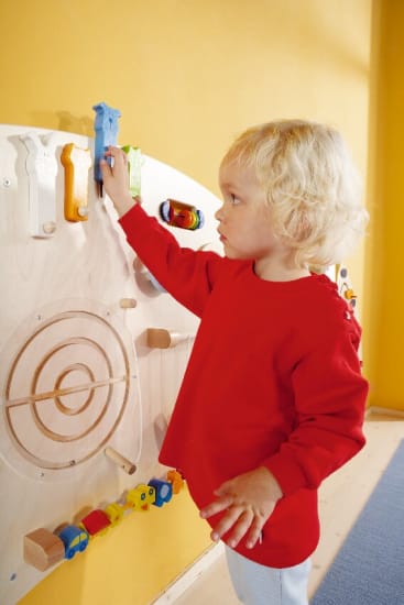Ein kleines Kind mit blondem Haar und rotem Hemd steht an einer hölzernen Aktivitätswand, bewegt mit einer Hand eine blaue Figur und berührt mit der anderen das Brett. Die Wand besteht aus verschiedenen bunten Holzelementen.