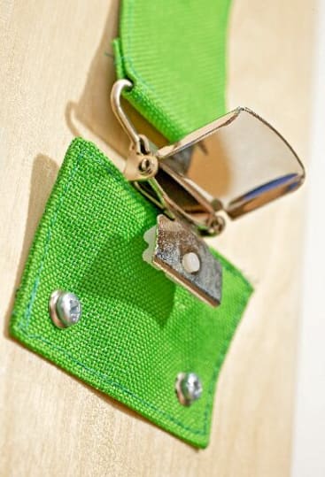 Nahaufnahme einer glänzenden Metallschnalle, die an einem hellgrünen Stoffband befestigt ist, das mit zwei silbernen Schrauben auf einer Holzplatte befestigt ist.