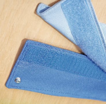 Nahaufnahme von zwei sich überlappenden blauen Stoffbändern mit Klettverschlüssen, die auf einer hellen Holzoberfläche liegen. Ein Band hat am Ende einen Druckknopf aus Metall.