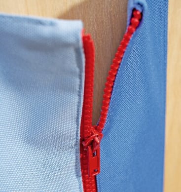 Nahaufnahme eines roten Reißverschlusses, der zwischen zwei blauen Stoffstücken teilweise geöffnet wurde, wobei im Hintergrund ein Teil einer hellbraunen Holzoberfläche zu sehen ist.