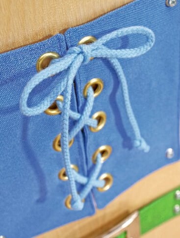 Nahaufnahme eines blauen Stoffes mit goldenen Ösen, der mit einem hellblauen Schnürsenkel zusammengeschnürt und mit einer Schleife versehen ist. Der Hintergrund scheint eine Holzoberfläche zu sein.