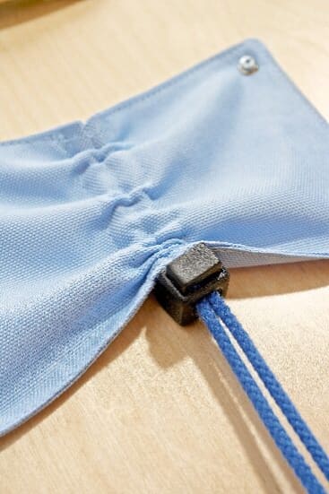 Nahaufnahme eines blauen Stoffes mit einer verstellbaren elastischen Schnur, die durch ein schwarzes Kunststoffschloss gezogen ist und auf einer hellen Holzfläche liegt.