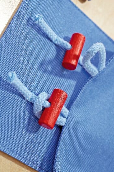 Nahaufnahme von zwei blauen Stoffschlaufen, die mit hellblauen Seilen und roten zylindrischen Holzknebeln auf einer blauen Stofffläche befestigt sind.