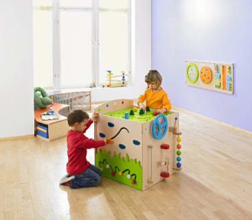 Zwei kleine Kinder spielen mit einem großen Aktivitätswürfel aus Holz in einem hellen, geräumigen Raum mit hellen Holzböden, weißen Wänden und Lernspielzeug. Das Sonnenlicht fällt durch ein Fenster im Hintergrund.