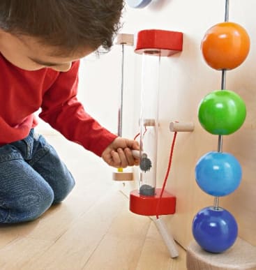 Ein Kind kniet auf einem Holzboden und interagiert mit einer sensorischen Spielwand, die eine durchsichtige vertikale Röhre mit kleinen schwarzen Objekten darin und eine vertikale Stange mit bunten Holzkugeln enthält.