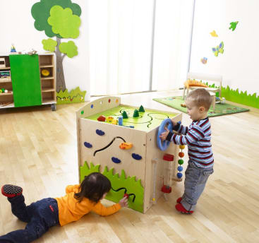 Zwei kleine Kinder spielen mit einem Aktivitätswürfel aus Holz in einem hellen, geräumigen Raum mit Holzböden, grünen Dekorationen und Spielzeug. Ein Kind steht, das andere liegt auf dem Boden, beide beschäftigen sich mit verschiedenen Seiten des Würfels.