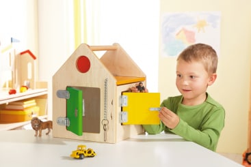 Ein kleines Kind öffnet eine gelbe Riegeltür an einem hölzernen Spielhaus mit mehreren Türen und Schlössern. Spielzeugfiguren, ein Löwe und ein gelber Lastwagen stehen auf dem Tisch. An der Wand im Hintergrund hängen Zeichnungen.
