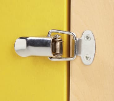 Nahaufnahme eines Metallriegels, der an der Kante einer gelben Holztür montiert und mit einer Metallplatte und Schrauben an einer hellen Holzoberfläche befestigt ist.