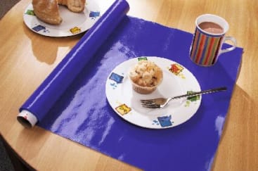 Auf einem runden Holztisch ist eine blaue, ausgerollte Matte teilweise ausgerollt. Auf der Matte stehen ein Teller mit einem Muffin und einer Gabel sowie ein gestreifter Becher mit einem Getränk. Im Hintergrund befindet sich ein weiterer Teller mit Brötchen.