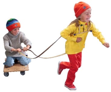 Ein junges Mädchen in einem gelben Hemd und einer roten Hose zieht ein jüngeres Kind mit Hilfe eines Geschirrs und Riemen auf einem Holzbrett mit Rädern. Beide Kinder tragen bunte Hüte. Der Hintergrund ist weiß.