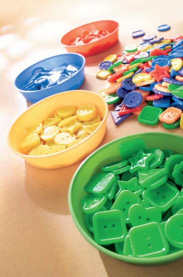 Vier Schalen mit verschiedenen bunten Kunststoffknöpfen in unterschiedlichen Formen. Die Knöpfe sind nach Farben sortiert: blau, rot, gelb und grün. Ein Stapel gemischter Knöpfe ist ebenfalls auf dem Tisch zu sehen.