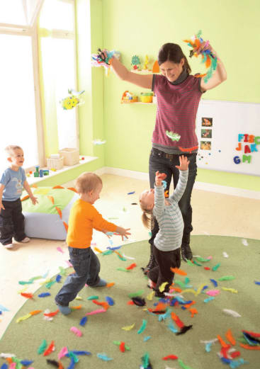 Ein Erwachsener und drei kleine Kinder spielen mit bunten Federn in einem hellen Raum. Die Federn sind auf dem Boden verstreut, und die Kinder greifen nach oben, während der Erwachsene die Federn in die Luft wirft.