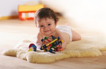 Ein Baby, das nur eine Windel trägt, liegt auf einem weichen cremefarbenen Teppich und hält ein buntes Spielzeug mit geometrischen Formen und eine Rassel in der Hand. Der Hintergrund ist sanft verschwommen und zeigt ein gelbes und oranges Objekt.