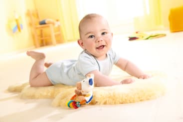 Ein Baby in einem hellen Strampler, das auf einem weichen, cremefarbenen Teppich in einem hellen Raum liegt, auf die Arme gestützt, mit einem Plüschhund und anderem Spielzeug im Hintergrund.