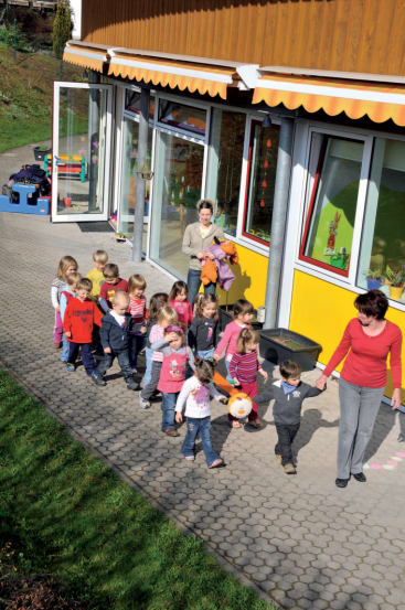 Eine Gruppe kleiner Kinder läuft in einer Reihe vor einer Kindertagesstätte oder einem Vorschulgebäude, beaufsichtigt von zwei Erwachsenen. Die Kinder halten sich an einem Seil fest und einige tragen einen Ball. Das Gebäude hat gelbe Wände und große Fenster.