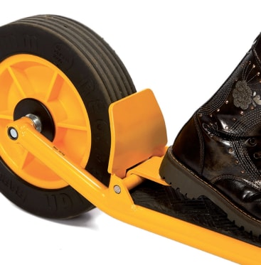 Nahaufnahme eines gelben Rollerrads und -rahmens mit einem schwarzen Stiefel, der auf der Fußplattform ruht. Das Bild hebt das Reifenprofil und einen Teil des Ziermusters des Stiefels hervor.